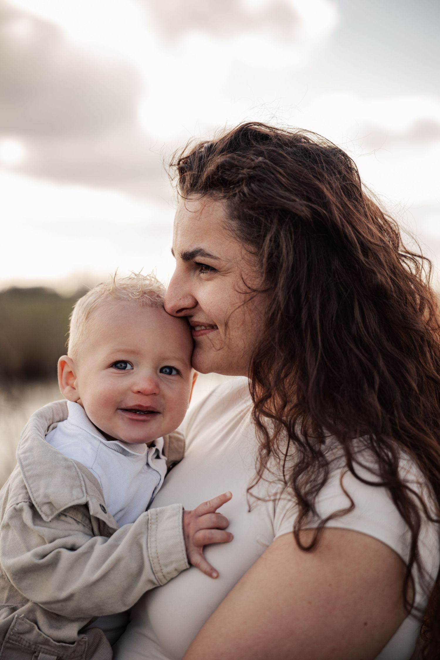 A curly-haired woman tenderly nuzzles a smiling baby boy outdoors under a cloudy sky.