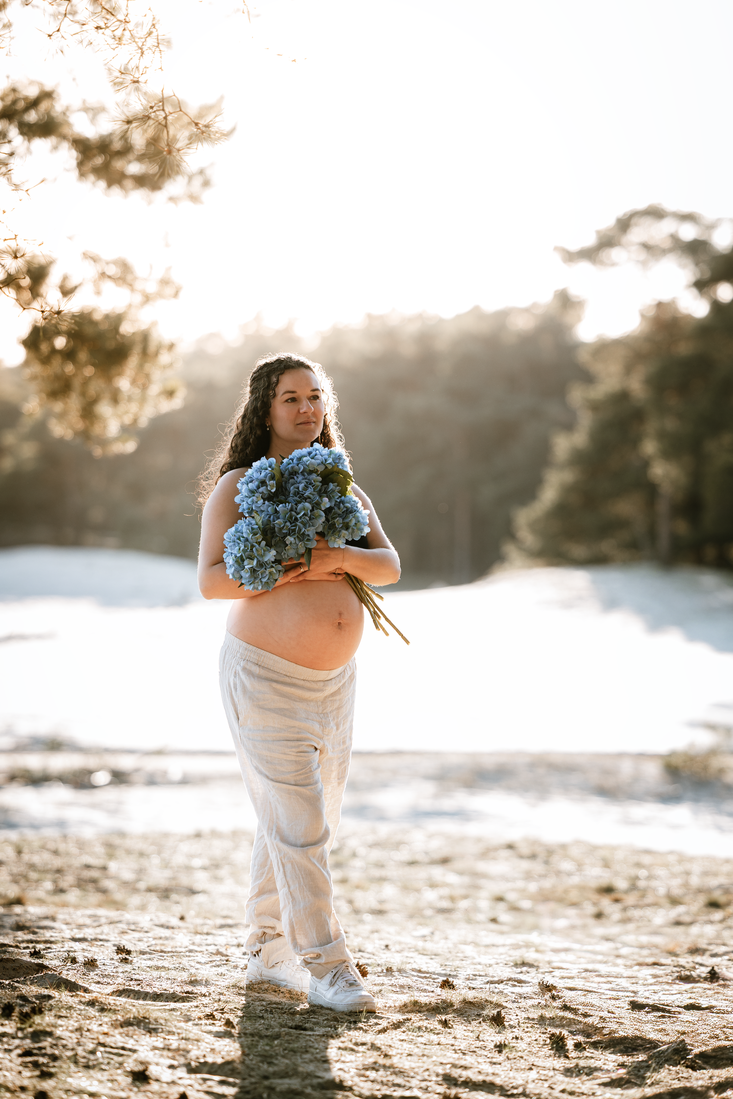 Expectant mother holds blue hydrangeas while standing on sandy beach at sunset in white outfit.
