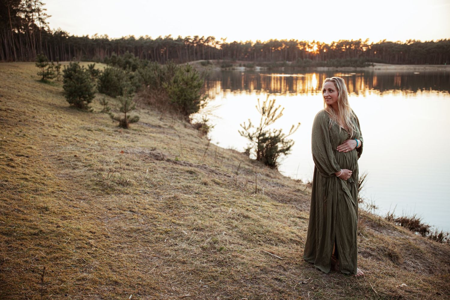Woman in green dress standing by lakeside during sunset in natural landscape.