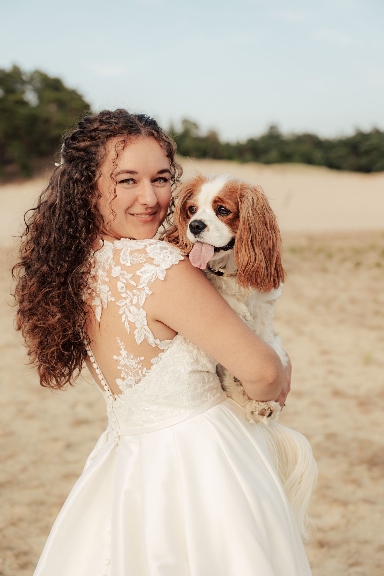 Cavalier King Charles Spaniel being held by someone in a lace wedding dress outdoors.