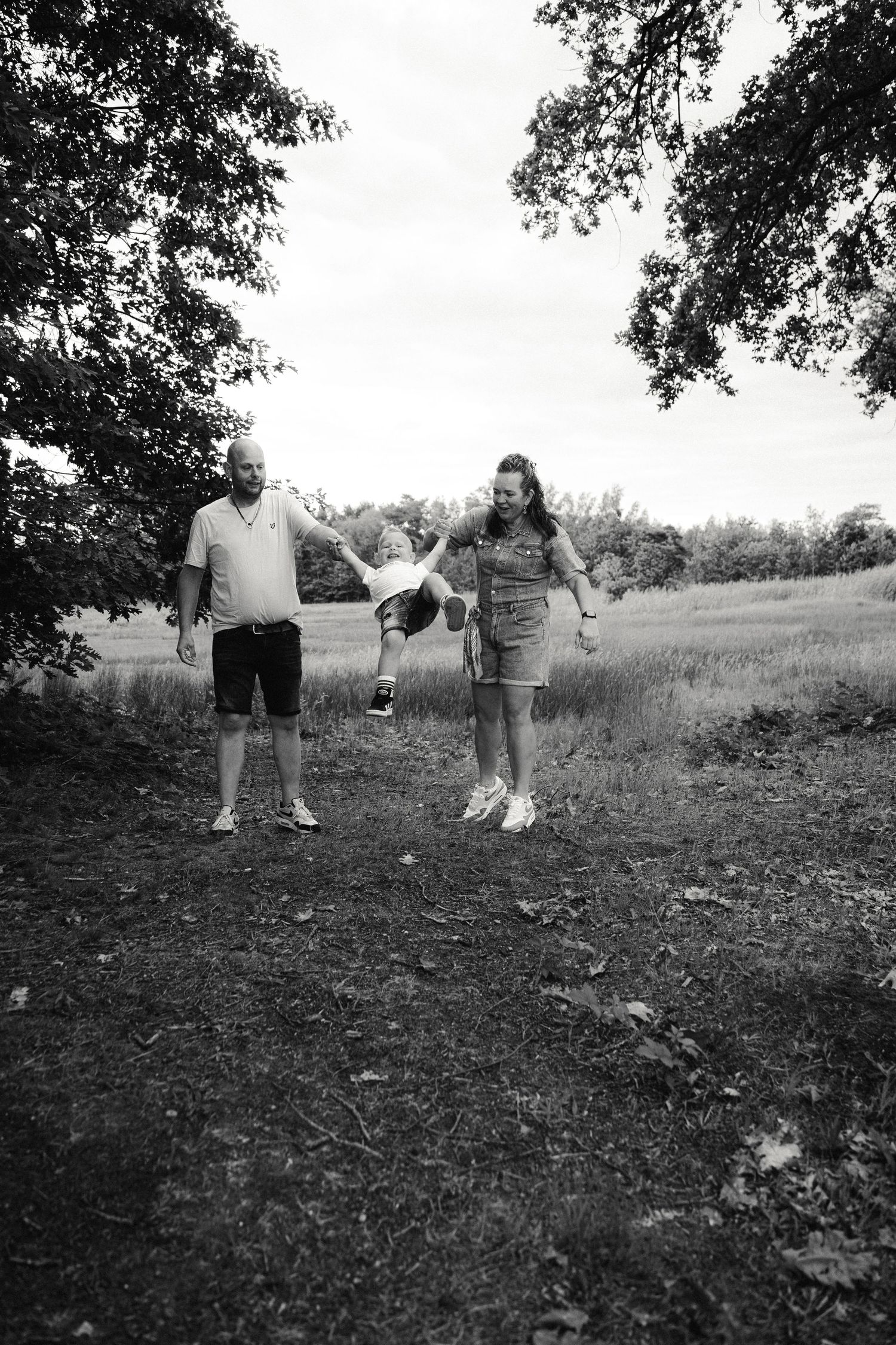 Black and white photo of parents swinging their child between them outdoors in a park setting.