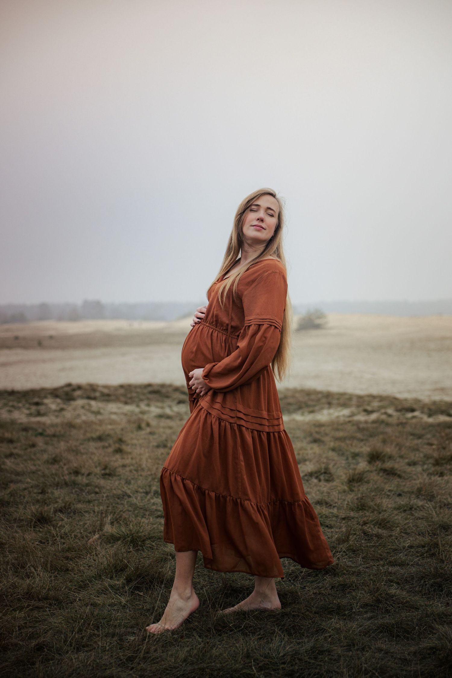 Woman in rust-colored dress poses for maternity photo in outdoor setting.