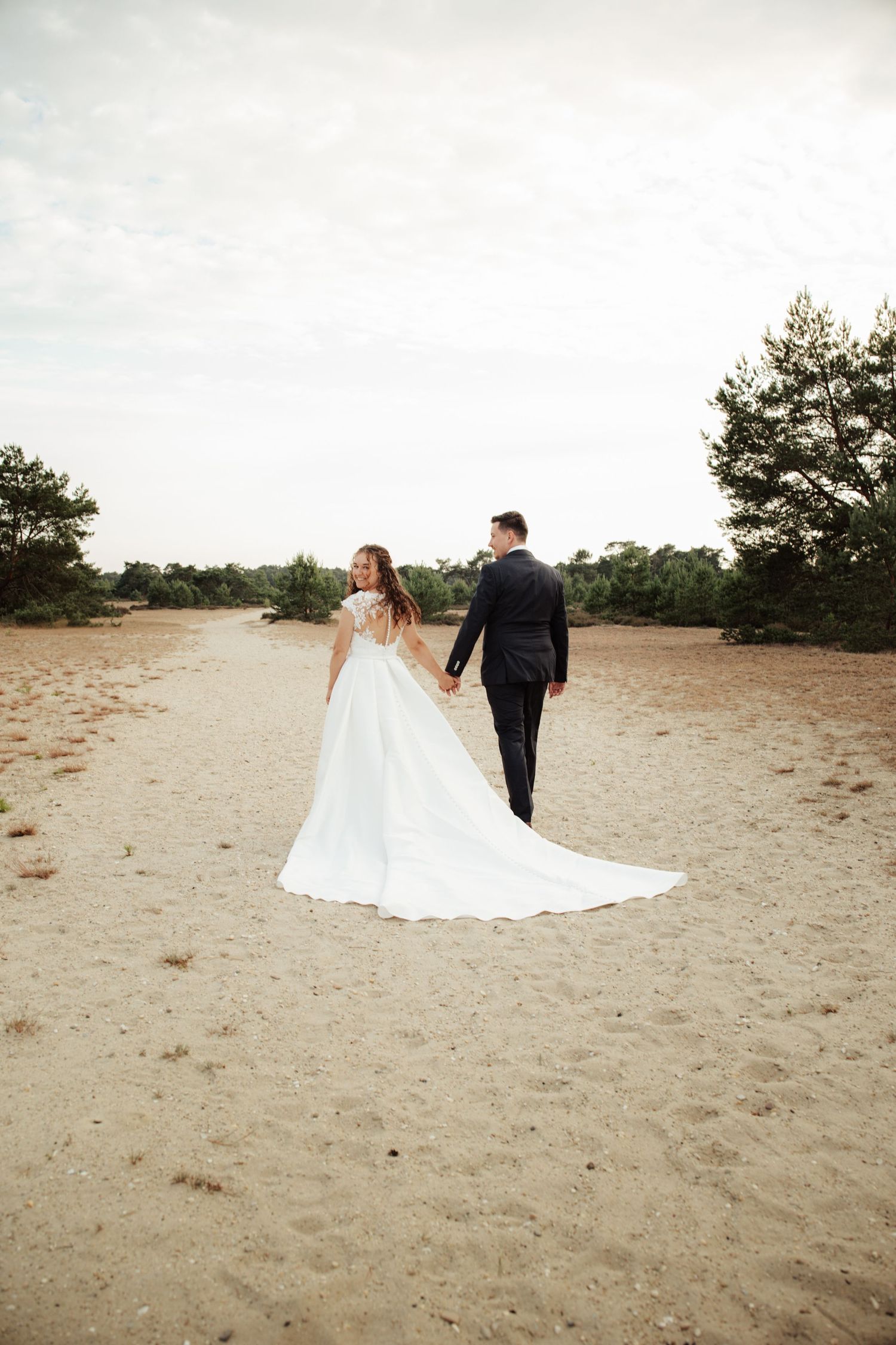 Bride and groom walking hand in hand on sandy path surrounded by trees in elegant wedding attire.