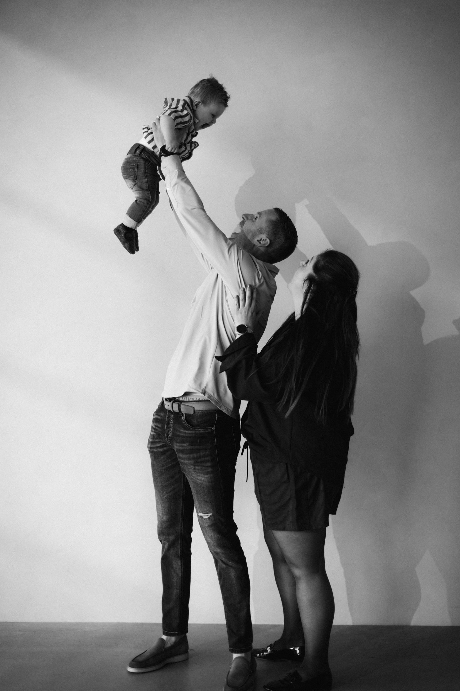 Black and white photo of a father lifting baby overhead while mother watches, family portrait against white wall.