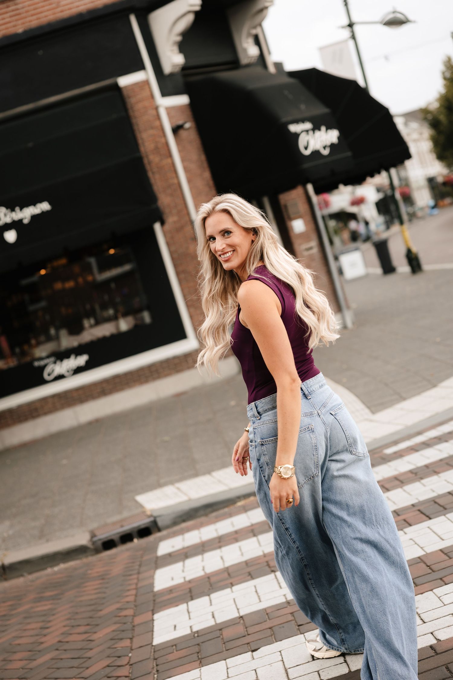 Blonde woman in purple top and wide-leg jeans posing on a crosswalk in an urban shopping district.