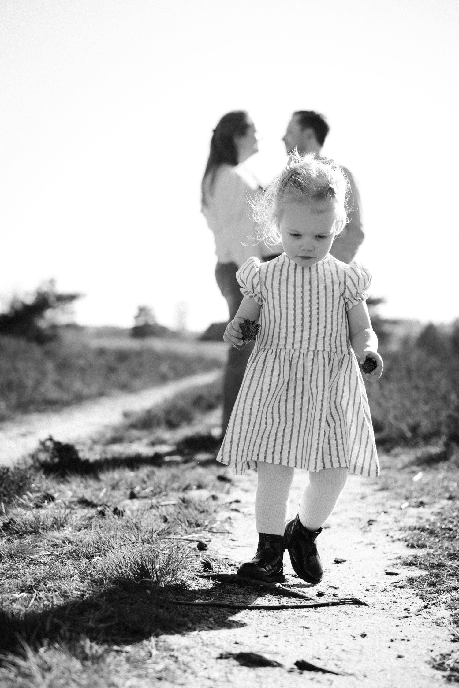 Black and white photo of a toddler girl in striped dress walking on a path while her parents stand together in background.