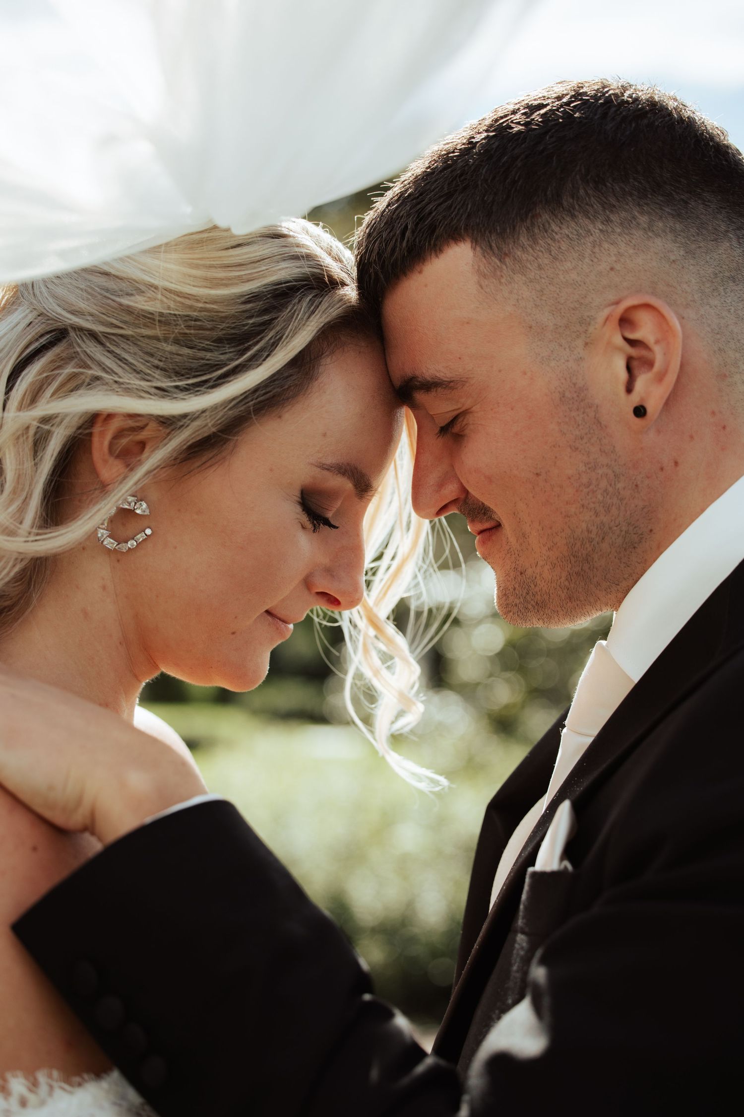A bride and groom share an intimate forehead touch moment, smiling softly under a flowing veil on their wedding day.