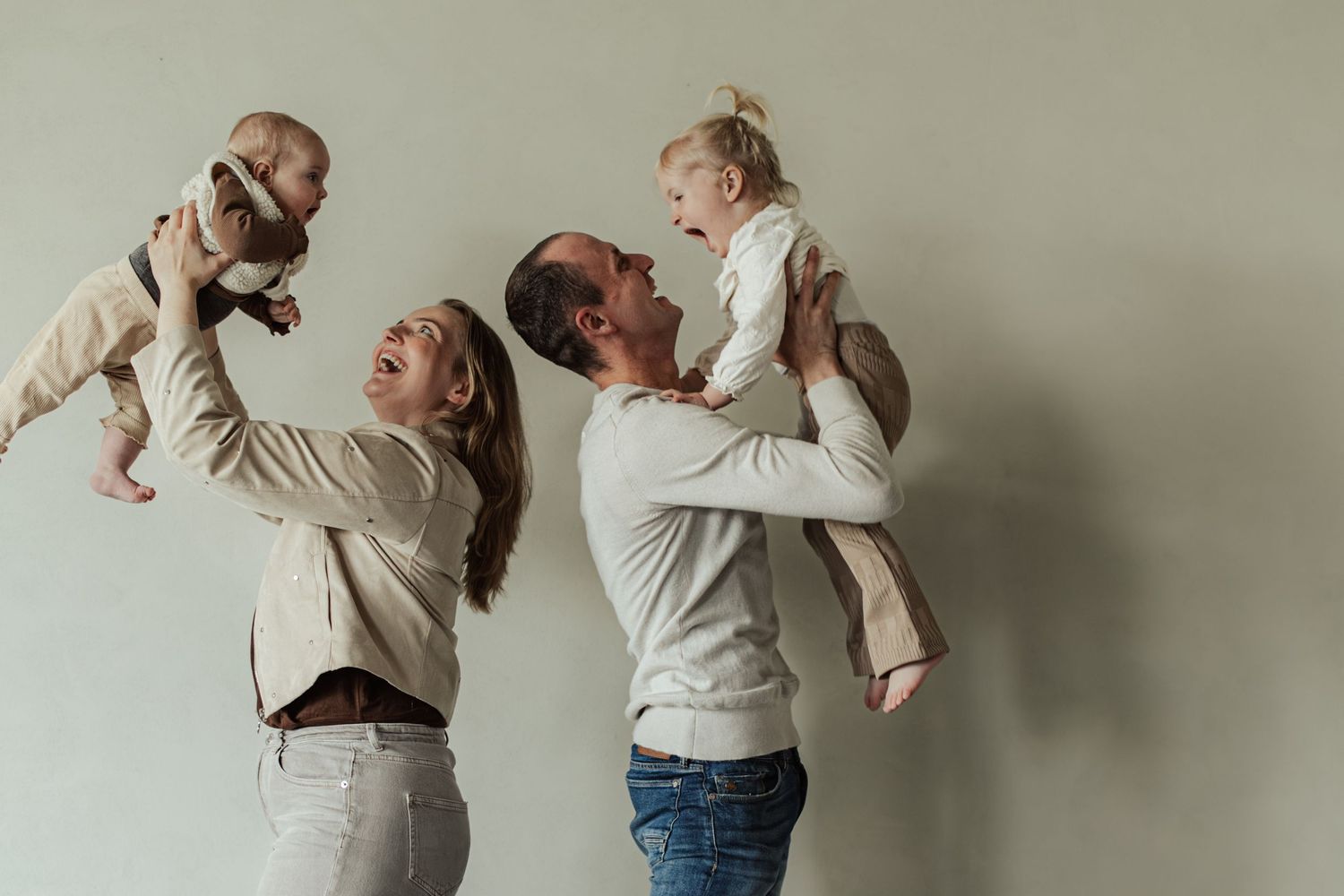Happy parents lifting two babies in the air, smiling joyfully against a light gray background.
