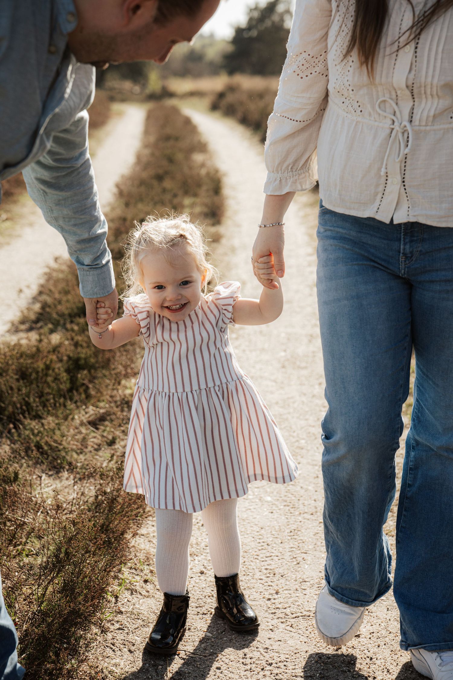 A smiling toddler girl in a striped dress holds her parents hands while walking on a sunny dirt path.