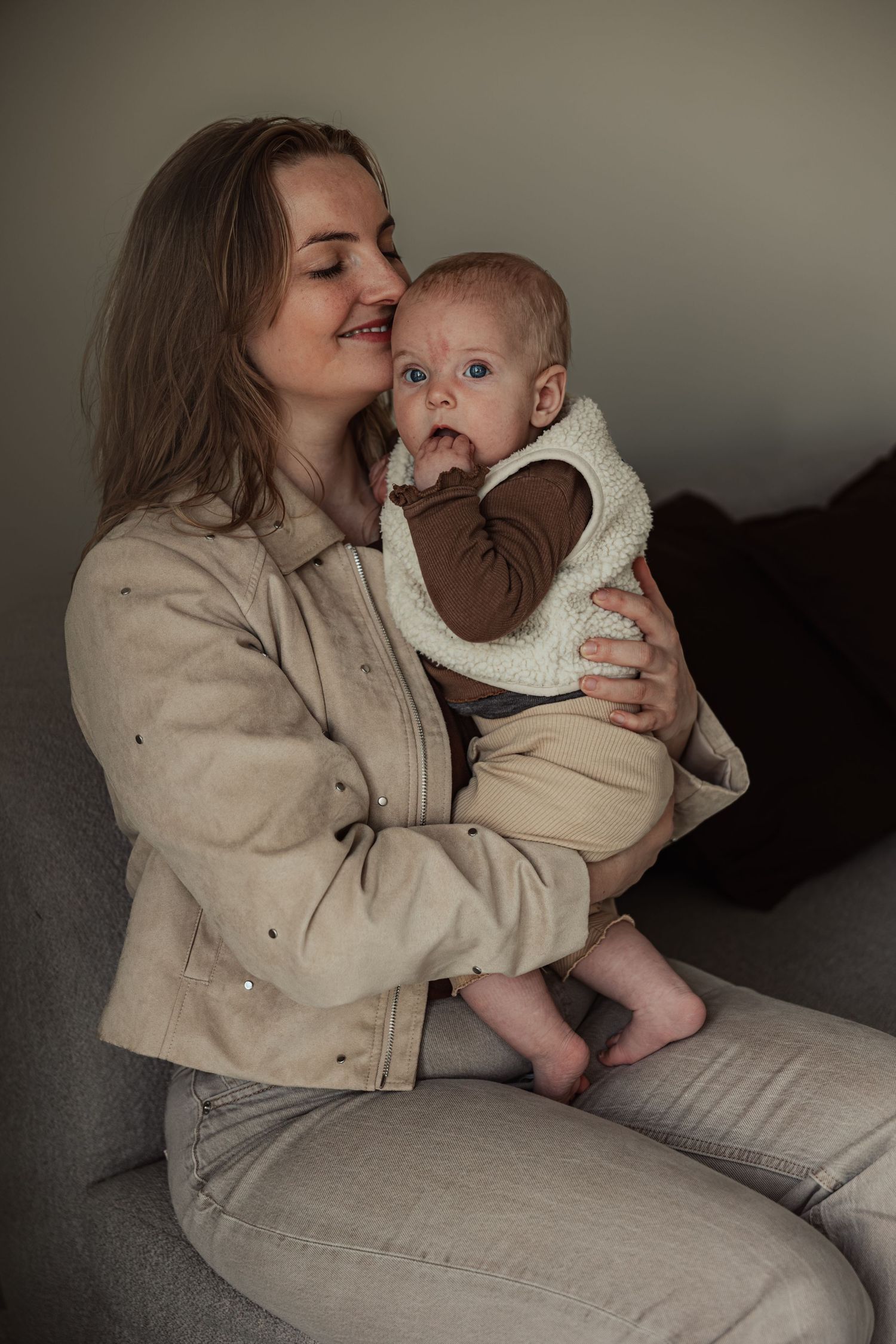 Young mother sitting on sofa holding her baby in sherpa vest, smiling lovingly in a warm home setting.