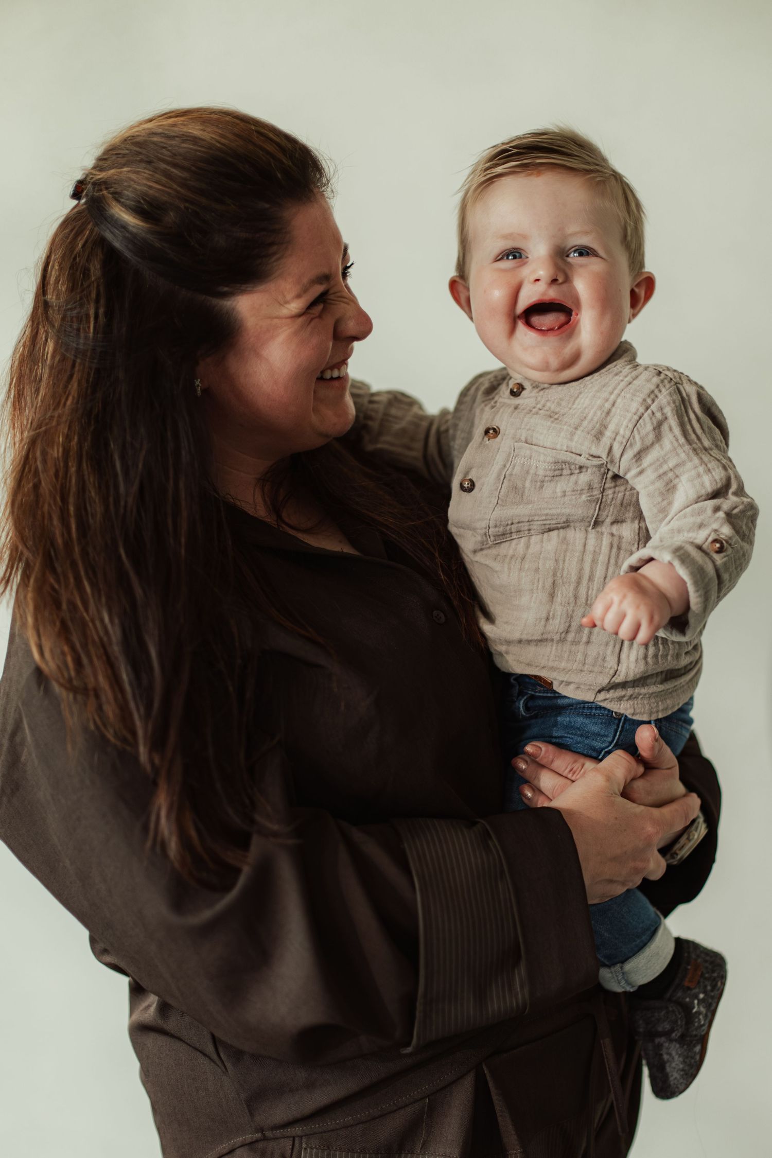 A smiling woman with long brown hair holds a laughing baby boy in a beige shirt and jeans against a white background.