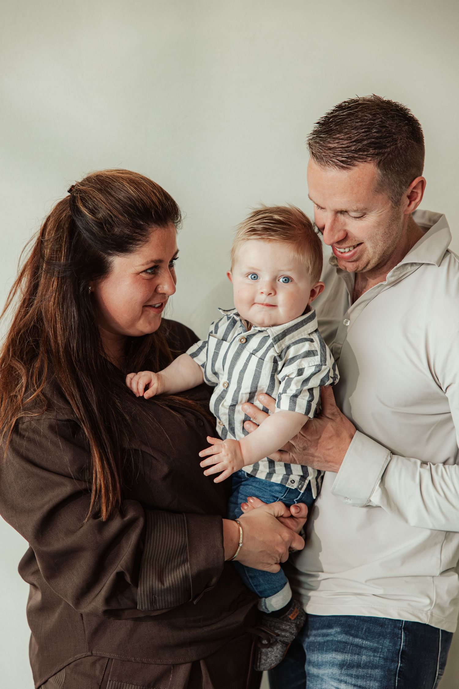 A young family with a baby boy in a striped outfit, parents smiling lovingly in a studio portrait.