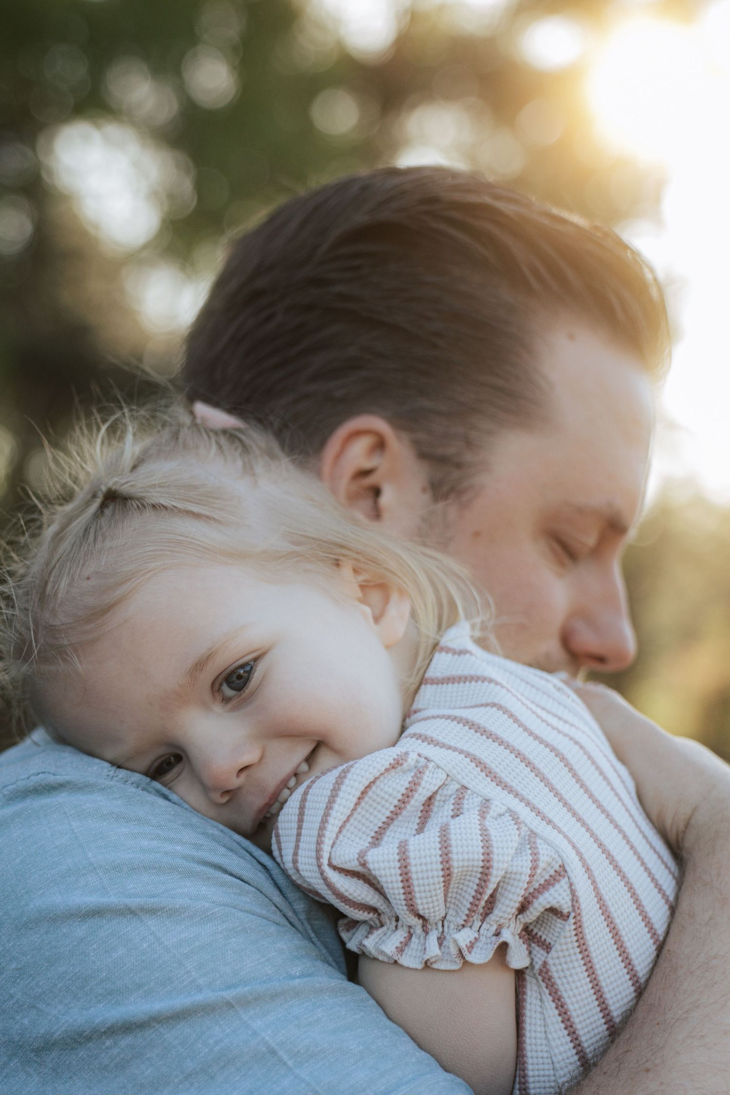 A young blonde toddler smiles while being held in a warm embrace by her father outdoors in golden sunlight.