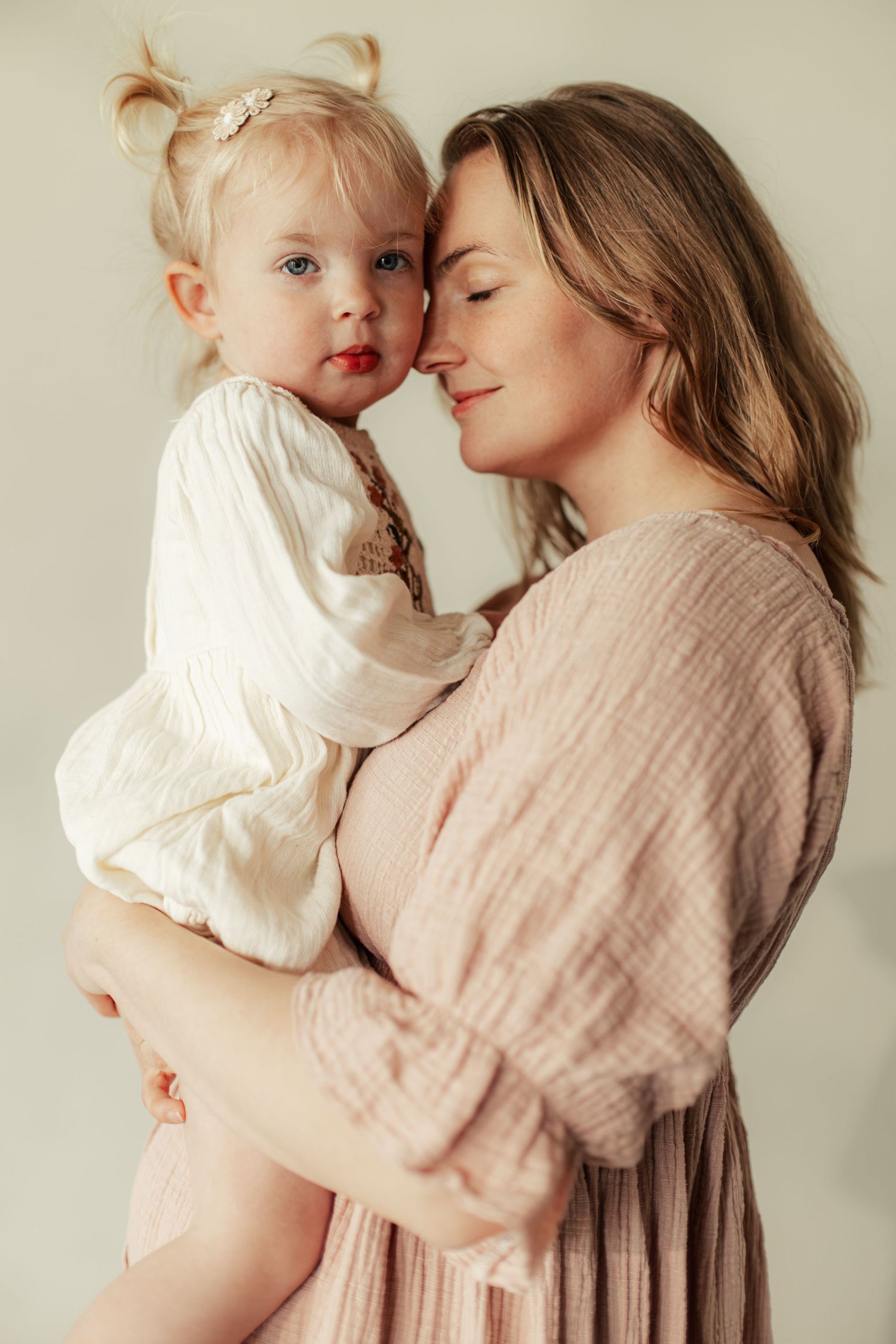 A tender moment as a mother in a pink dress holds her blonde toddler daughter, both in soft, neutral tones.