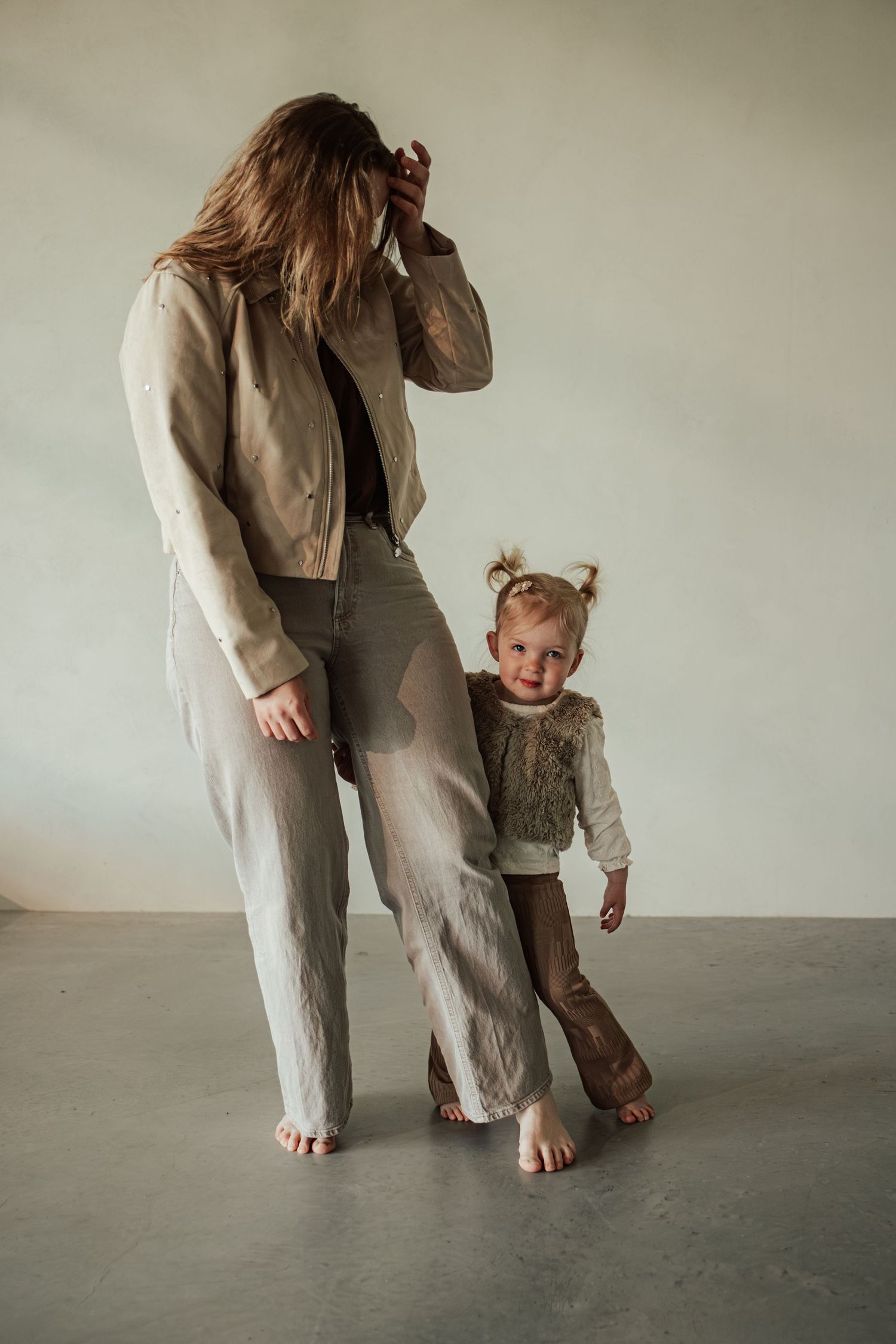 A woman in a beige jacket and jeans stands barefoot beside a toddler girl in a fur vest and brown pants.