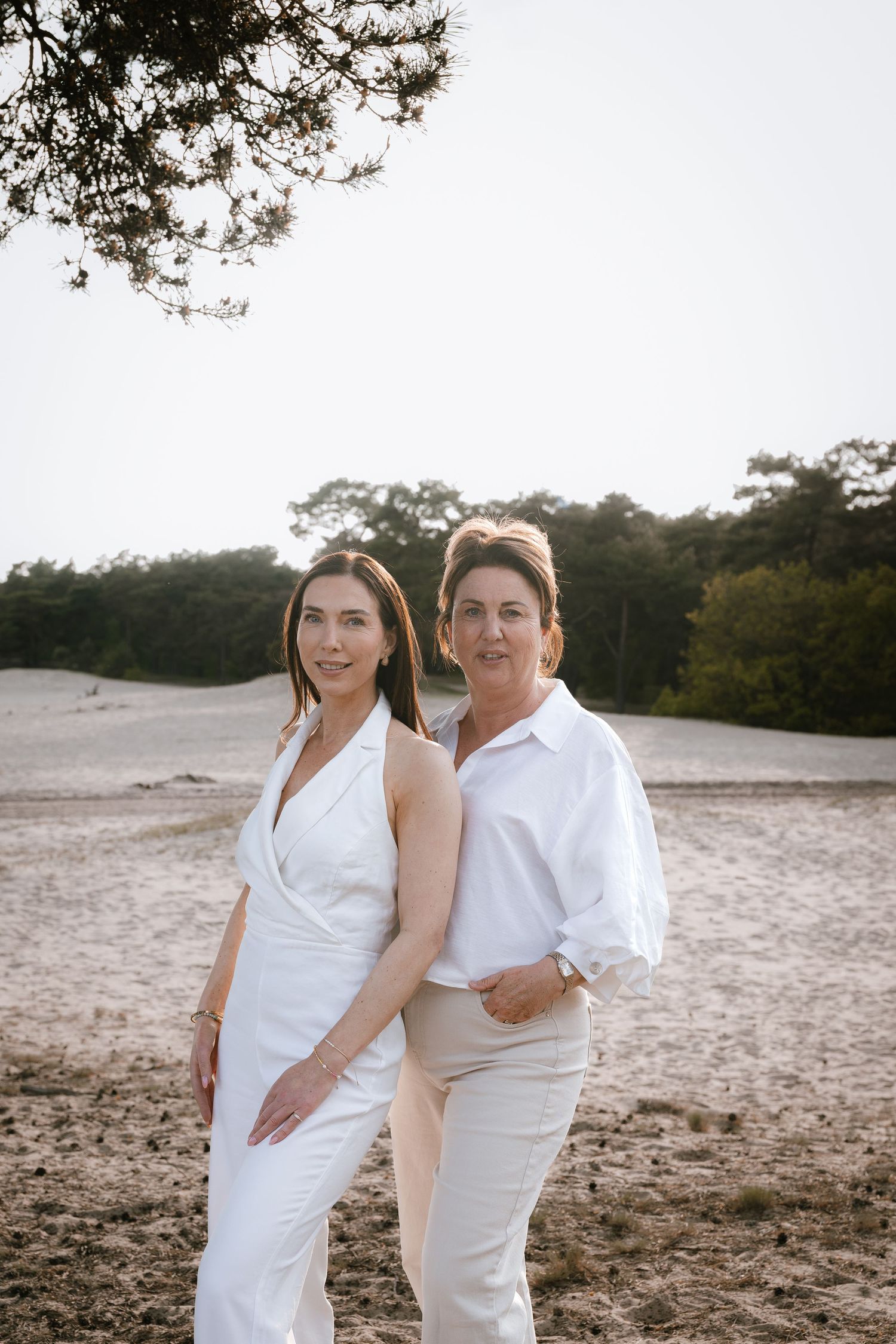 Two women in white outfits pose together on a sandy beach landscape with trees in the background.