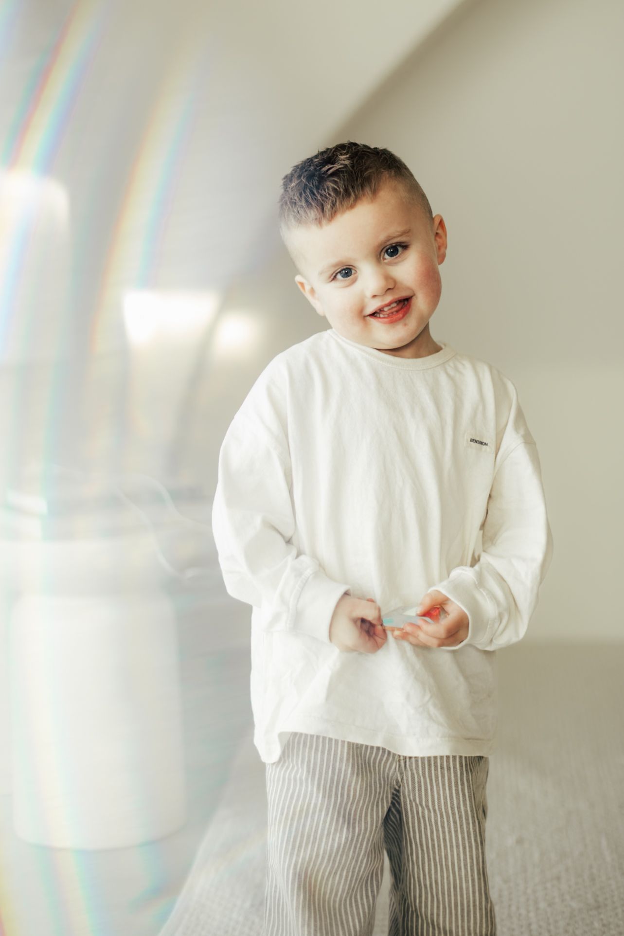 Young child in white sweater and striped pants smiling against white wall.