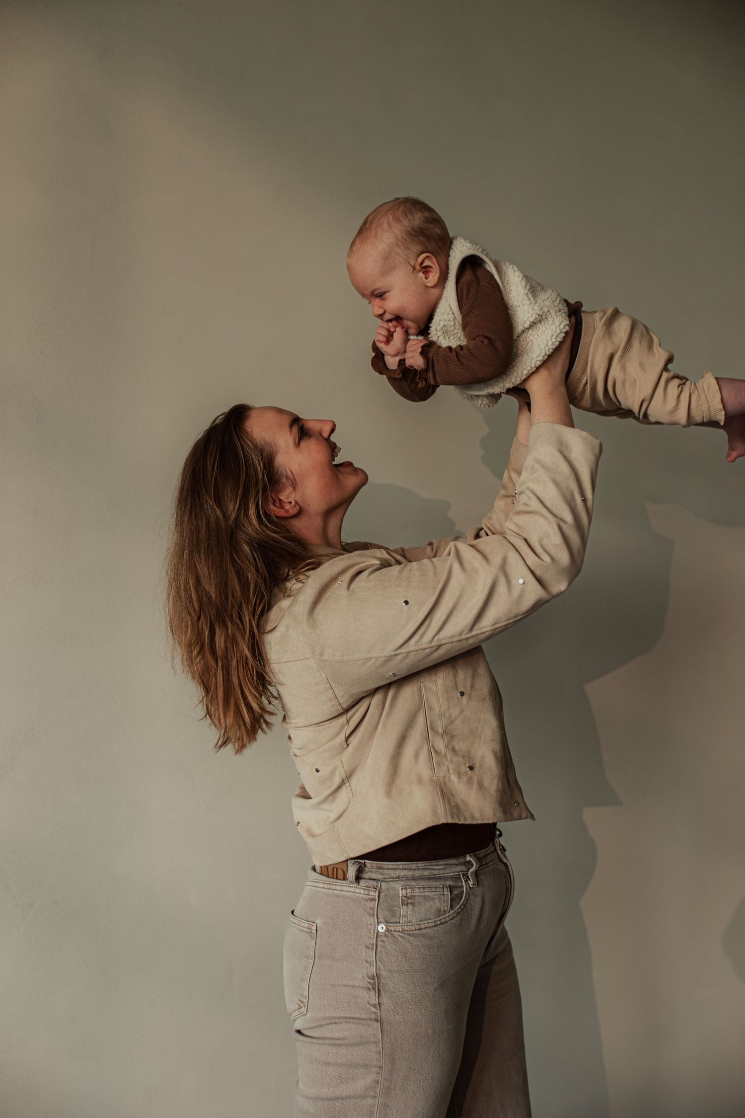 A joyful mother lifts her smiling baby overhead against a neutral background, both dressed in warm beige tones.