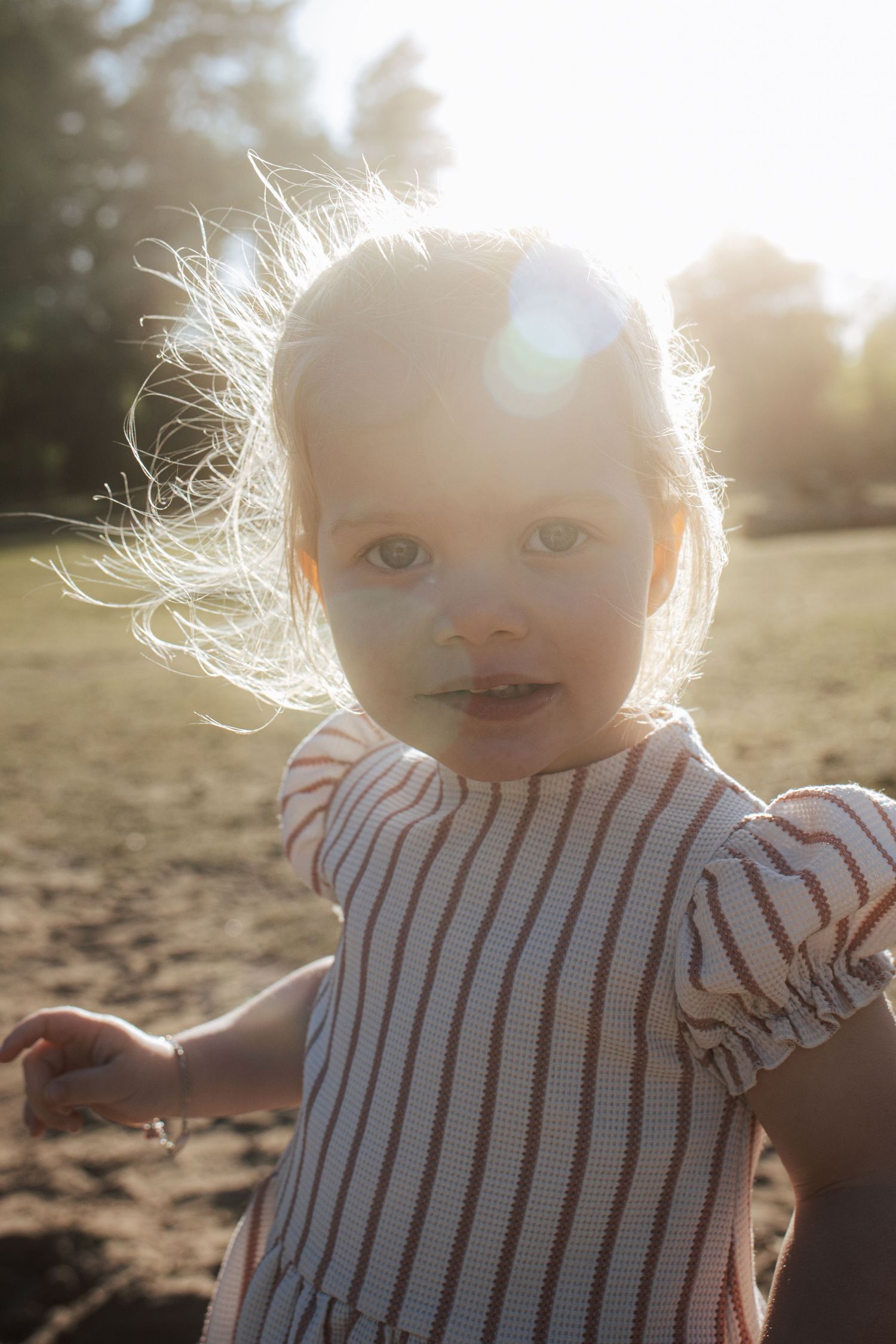 Toddler girl in striped dress outdoors in golden backlit sunlight with wind-blown hair.