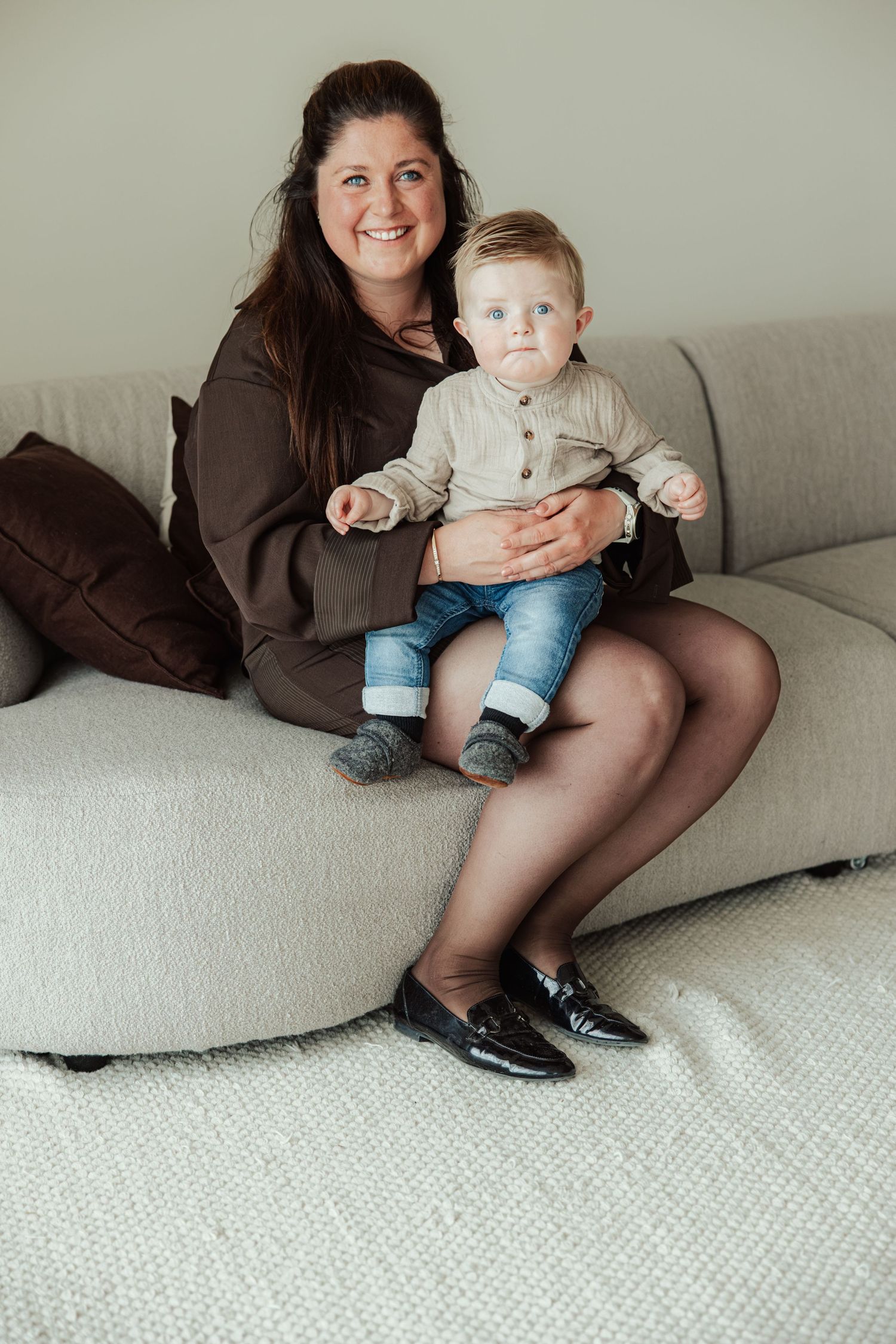 Smiling woman in brown dress holding a baby boy on a gray sofa at home.