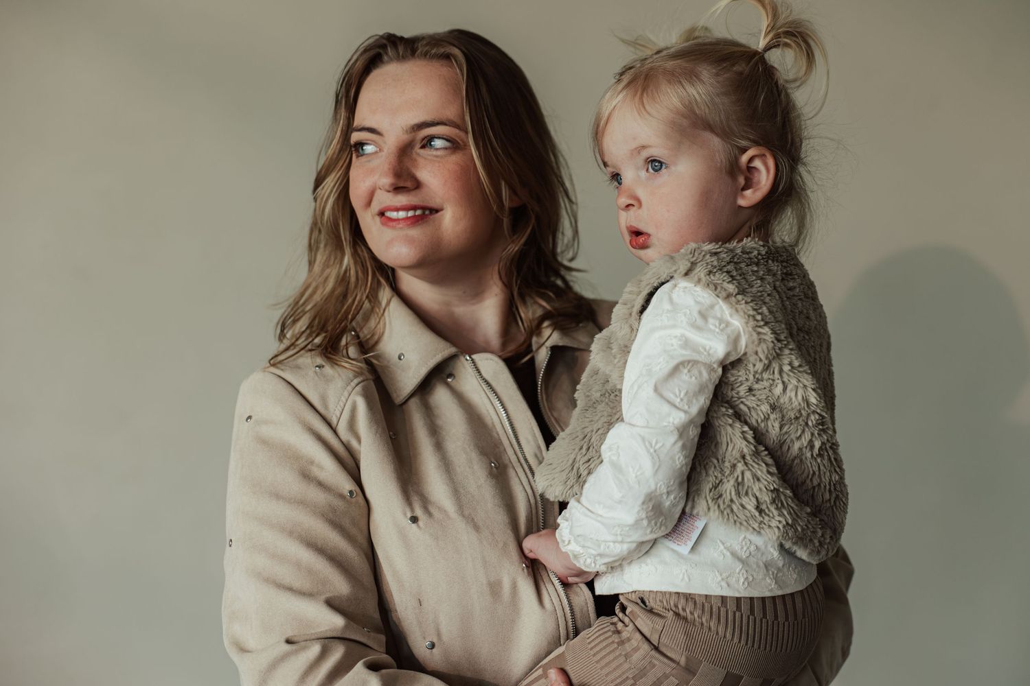 A woman in a beige coat holds a young blonde toddler girl in a knit vest against a soft gray background.