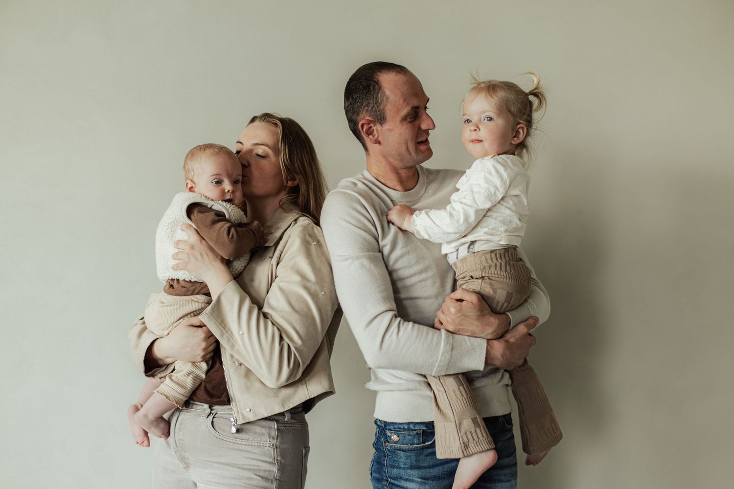 A happy family of four posing together, parents holding two young toddlers, smiling warmly against a neutral background.