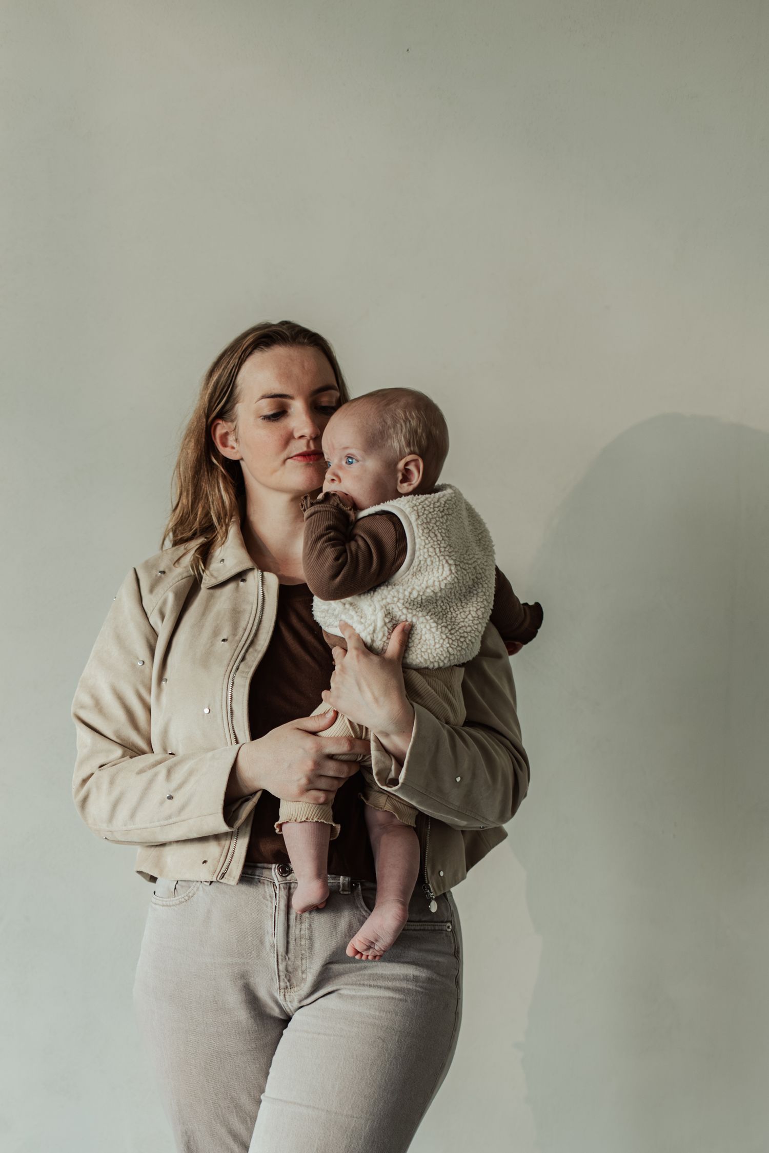 A young mother in a beige jacket tenderly holds her newborn baby against a soft white background.