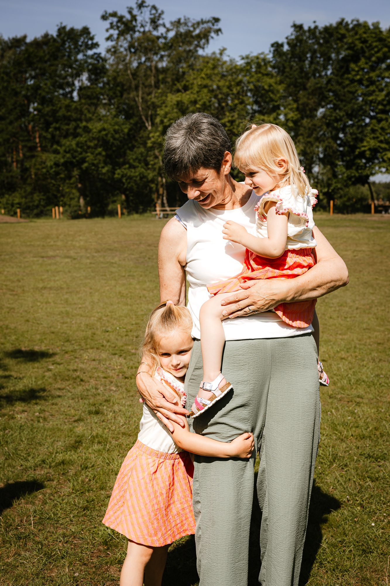 Grandmother holding toddler girl while another young girl hugs her waist outdoors in a sunny green park.