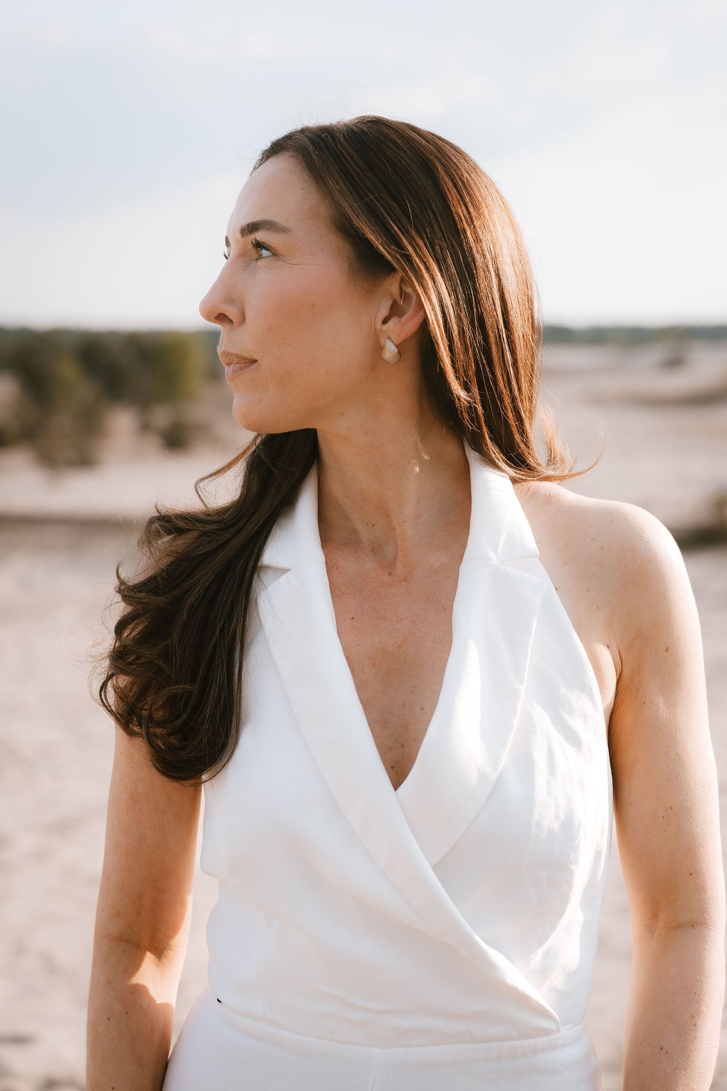 Woman in white halter dress standing on sandy beach, dark hair blowing, wearing pearl earrings.