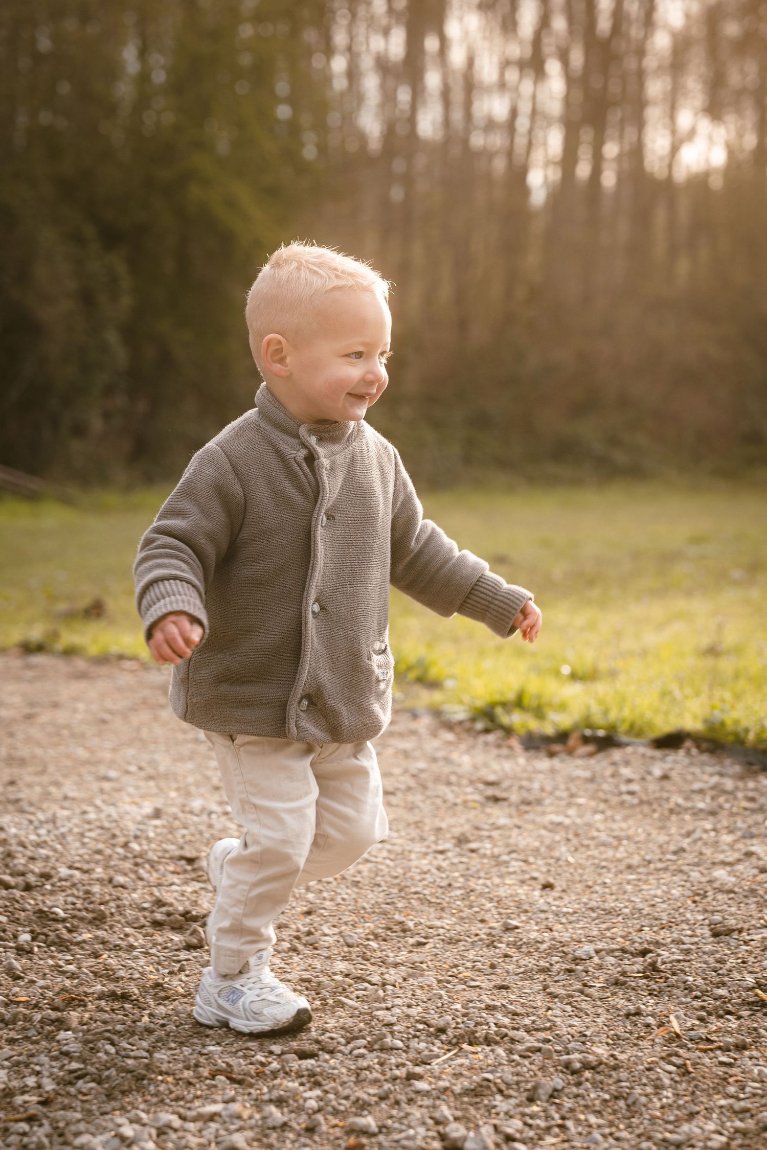 Smiling toddler boy in gray knit cardigan walking on gravel path in golden sunset light with trees in background.