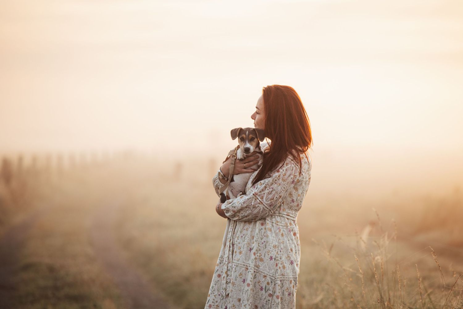 Someone in a floral dress holds a small dog while standing in a misty field at sunset.