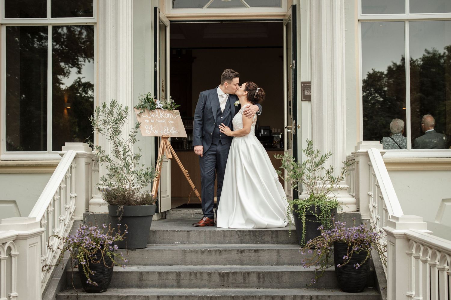 Elegant wedding couple stands on white porch steps decorated with potted plants.