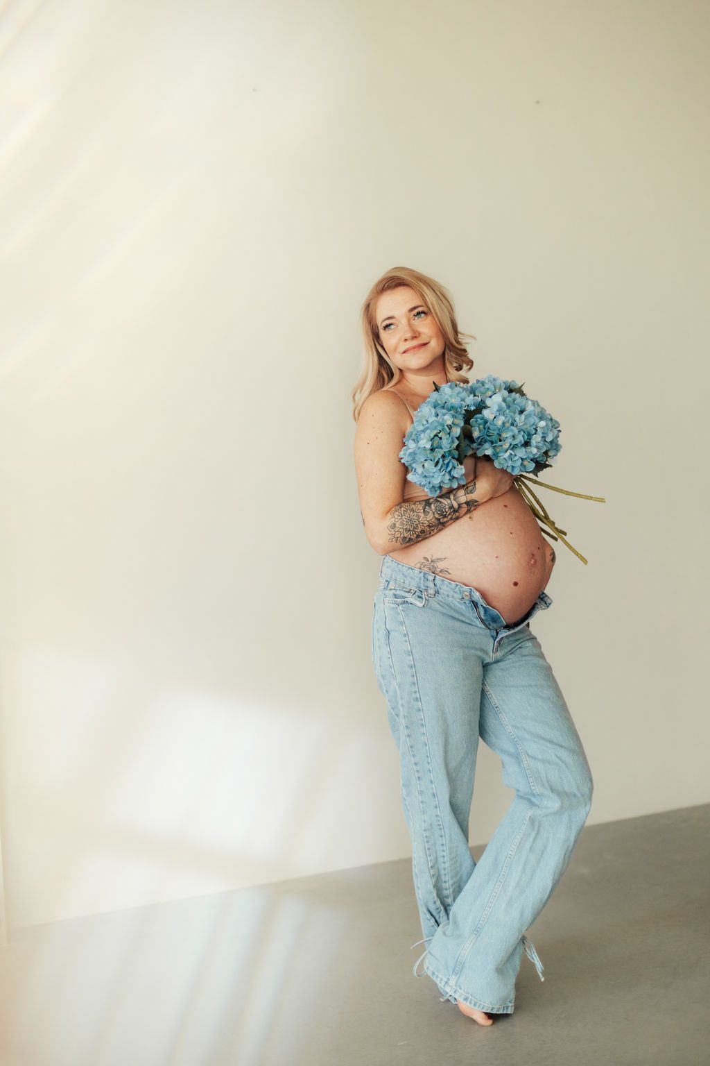 Maternity photo of an expectant mother in light jeans holding blue hydrangeas against a neutral wall.