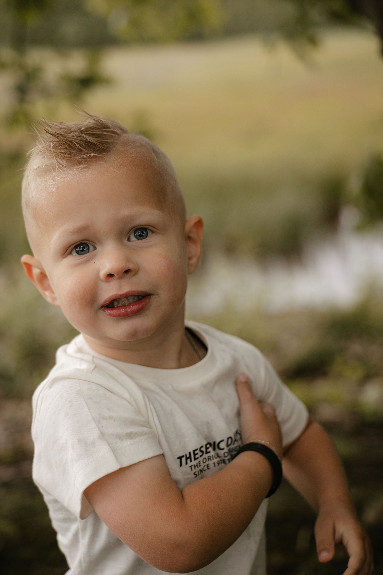 A young blonde toddler boy in a white graphic t-shirt smiles outdoors with a blurred green nature background.