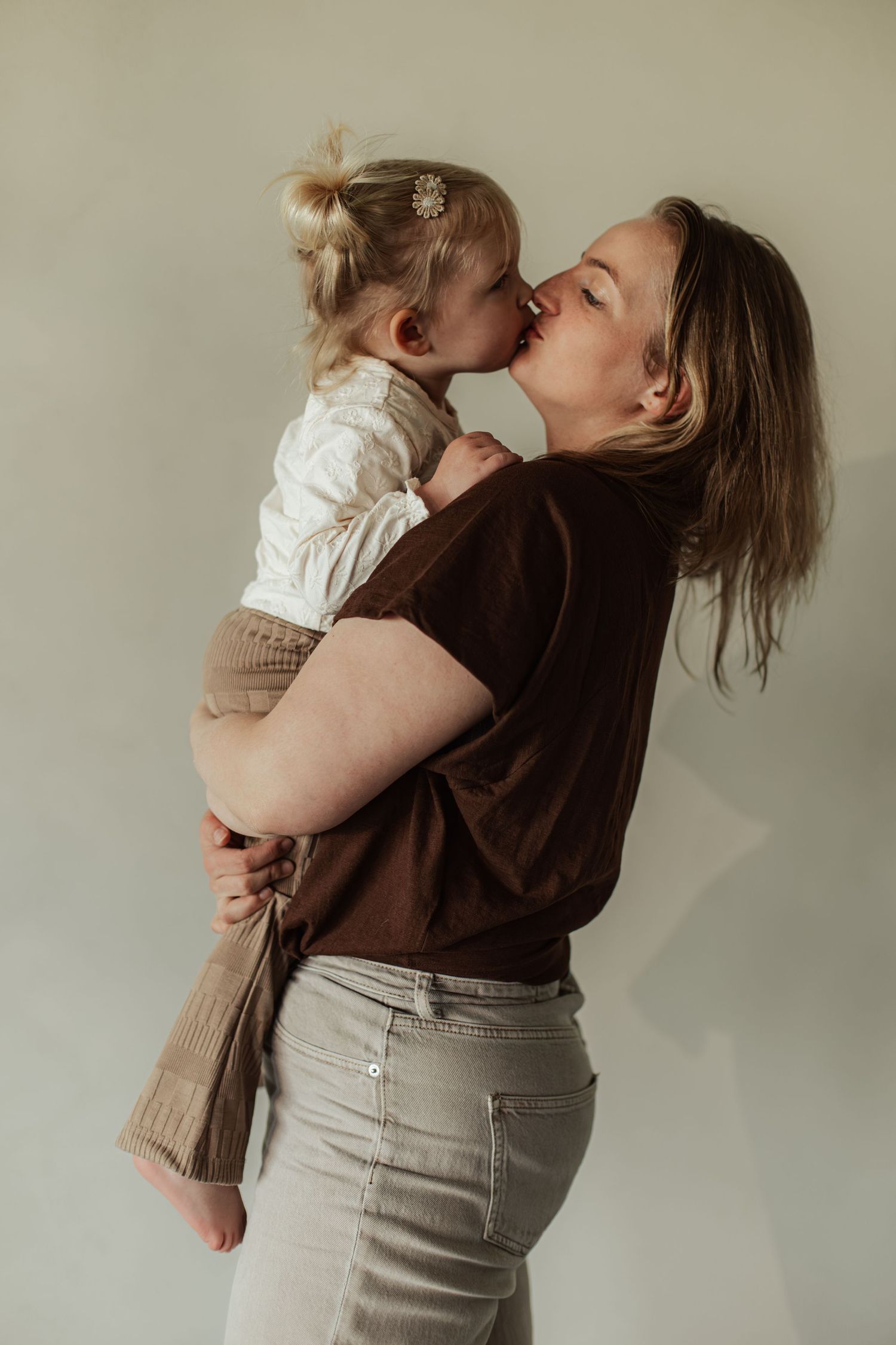 A mother tenderly kisses her toddler daughter while holding her against a neutral background.