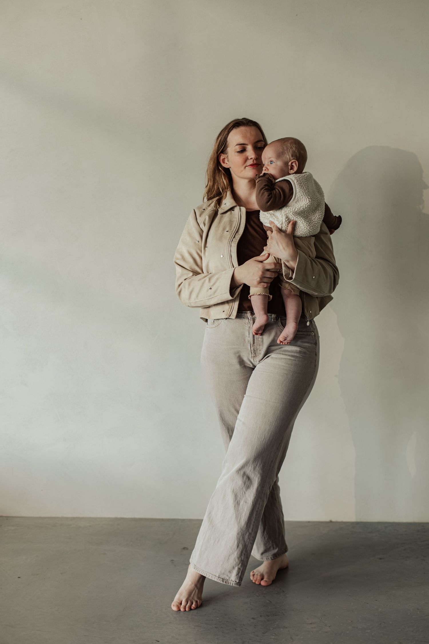 A young mother in beige wide-leg pants and jacket holds her newborn baby while standing barefoot against a light wall.