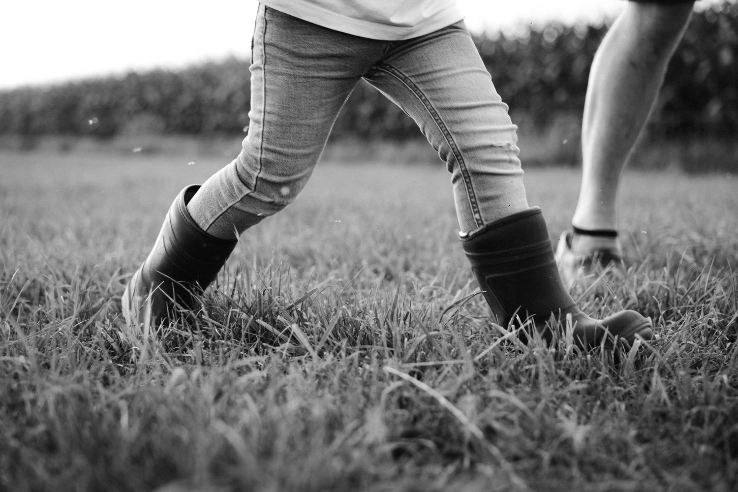 Child wearing jeans and rubber boots walking on grass in black and white photo.
