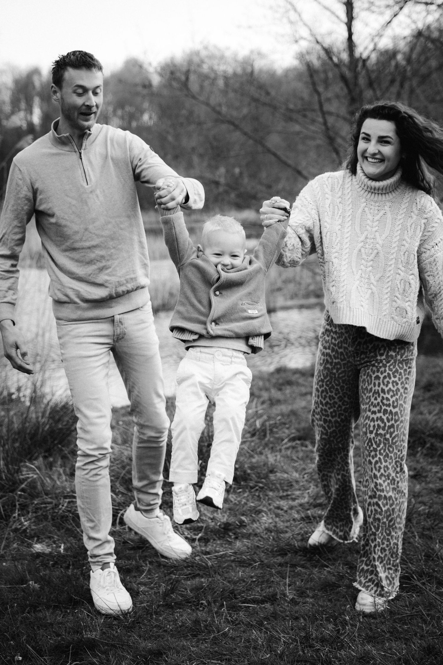 Black and white photo of a happy family swinging their toddler outdoors near a pond.