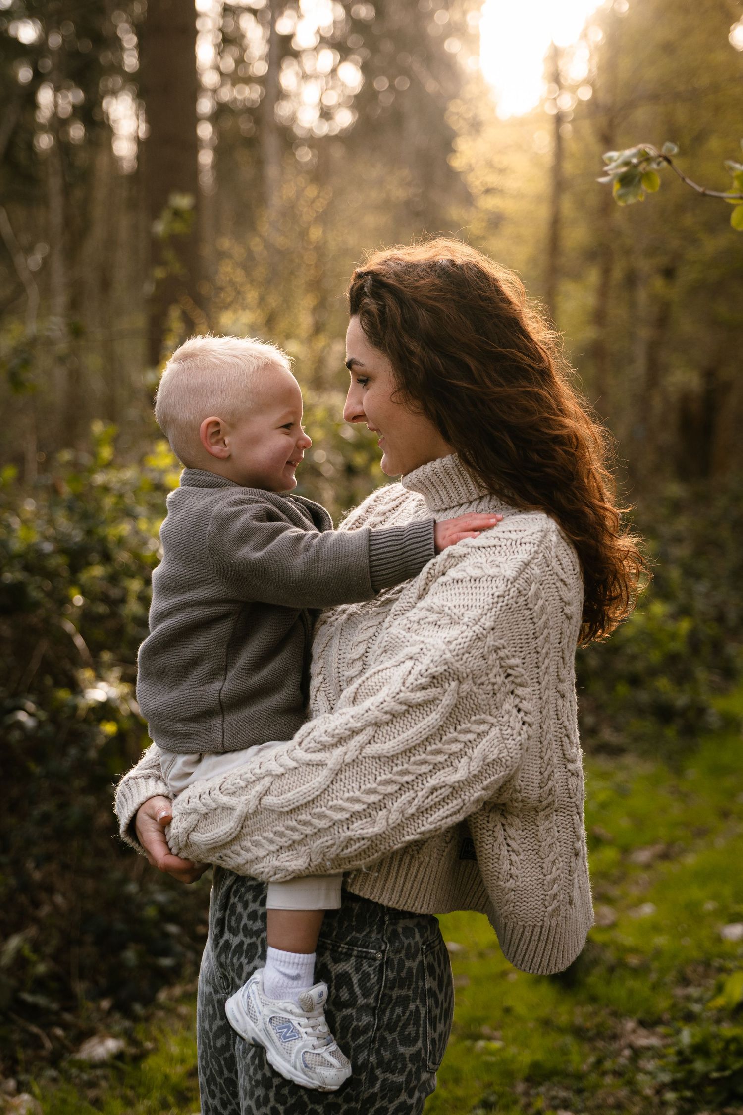 A mother holds her toddler in a sunlit forest, both smiling warmly, wearing cozy knit sweaters.