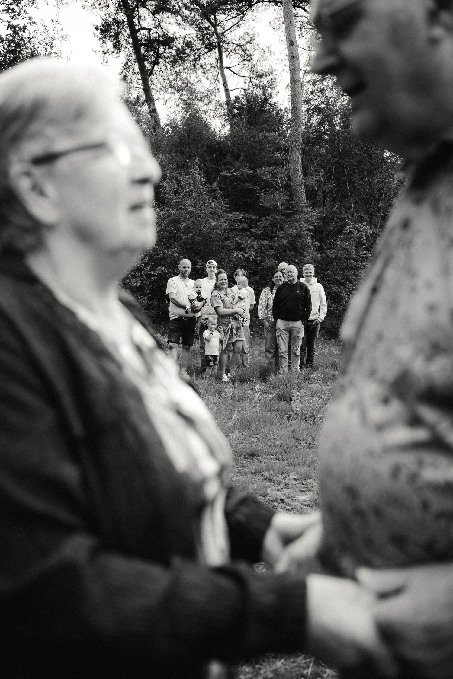 Black and white photo of a group of people standing in a forest clearing, framed by two blurred figures in the foreground.