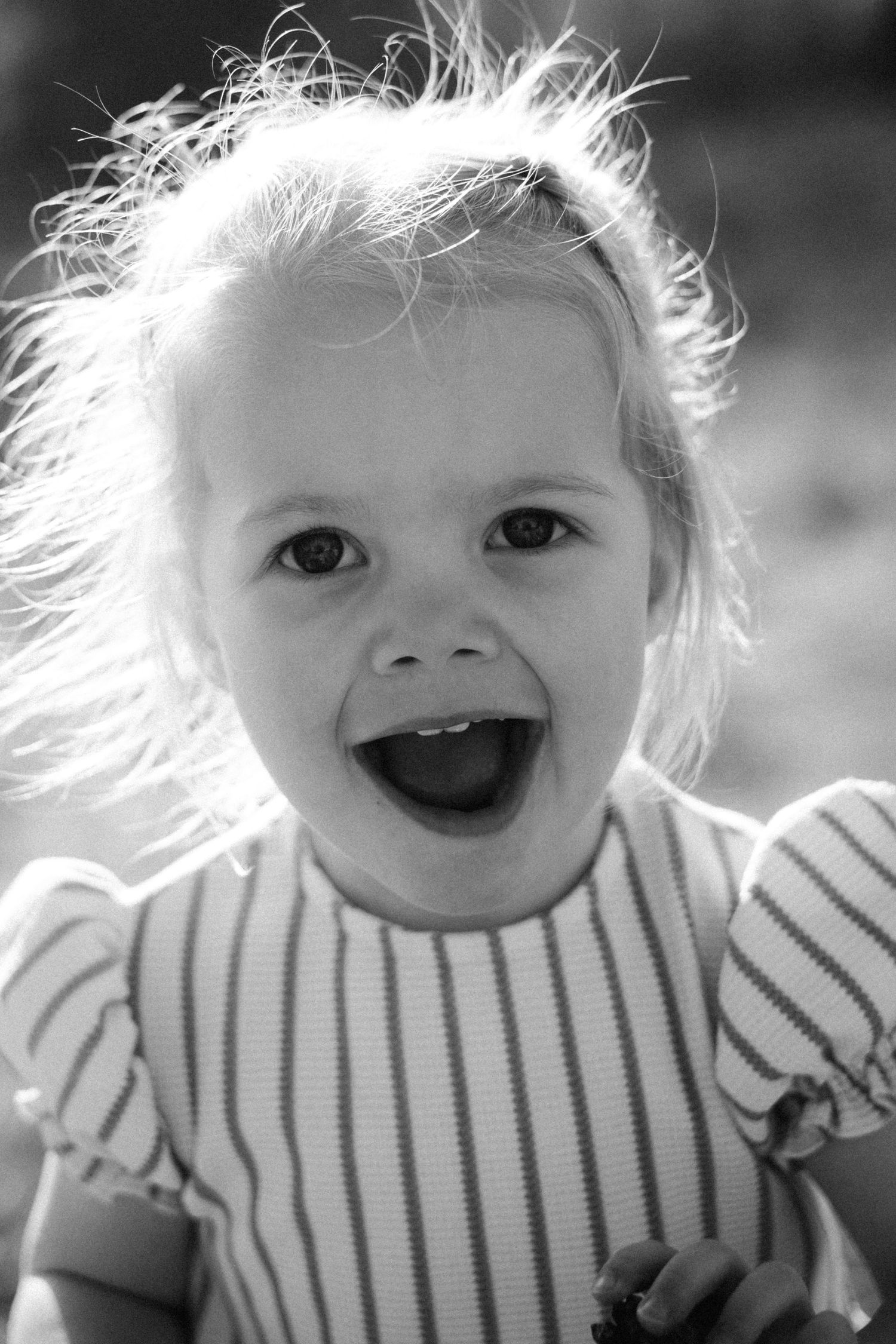 Black and white photo of a joyful toddler girl with windswept hair, laughing outdoors in a striped shirt.