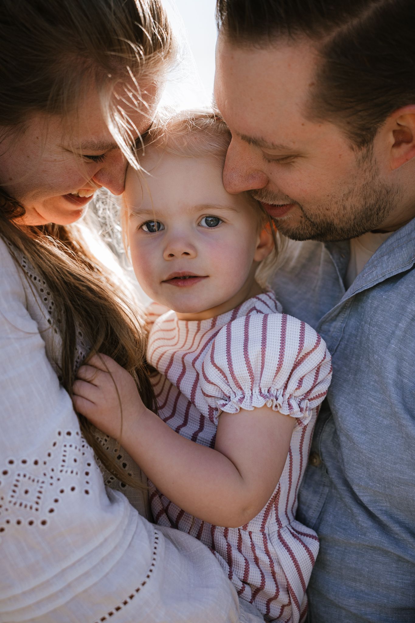 A young toddler girl in a striped dress cuddles between her loving parents outdoors in warm sunlight.