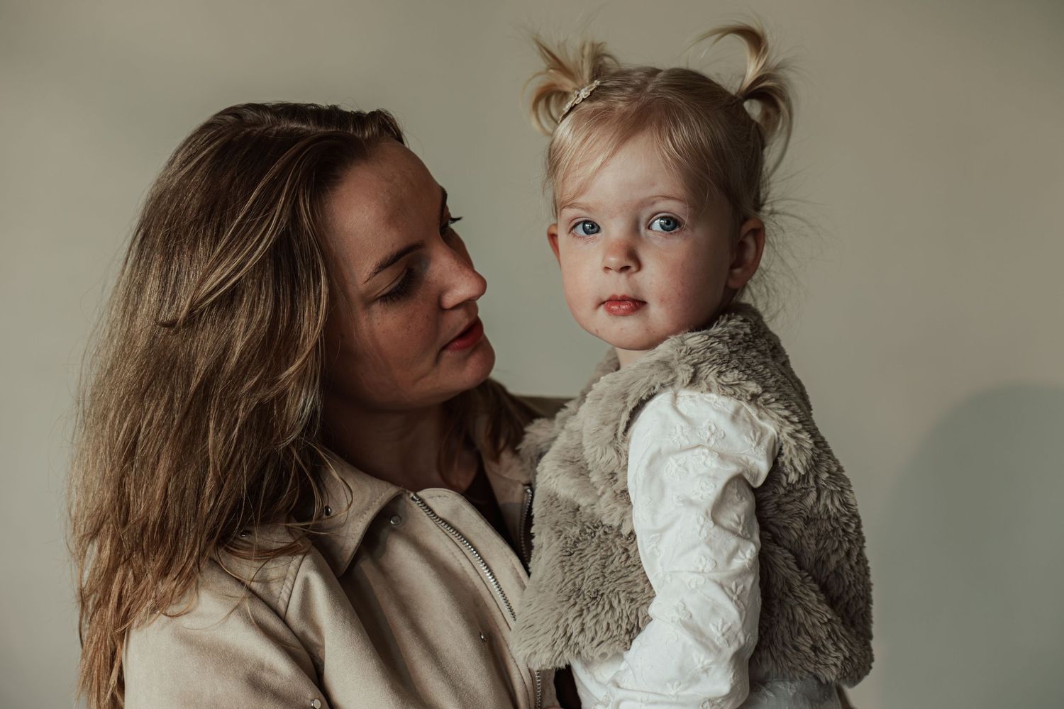 A mother tenderly holds her toddler daughter with pigtails, both sharing a gentle moment against a neutral background.