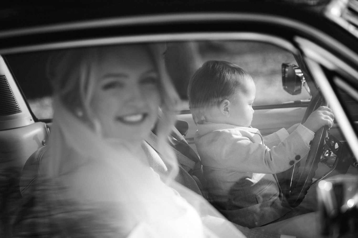 Black and white photo of a smiling bride sitting in a vintage car with a young child at the steering wheel.