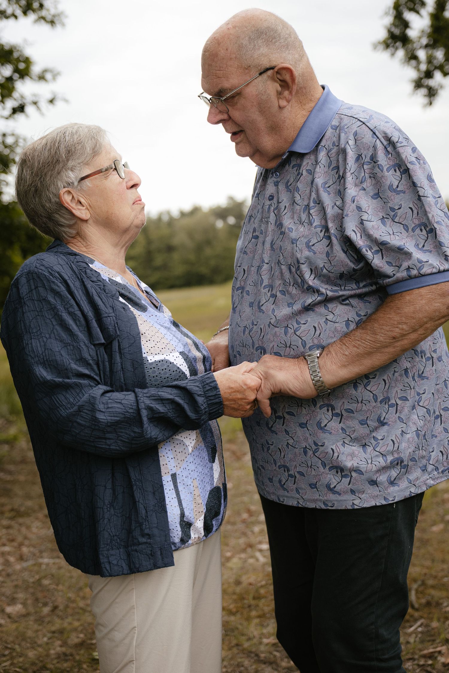 Elderly couple holding hands outdoors, sharing a tender moment surrounded by lush green trees.