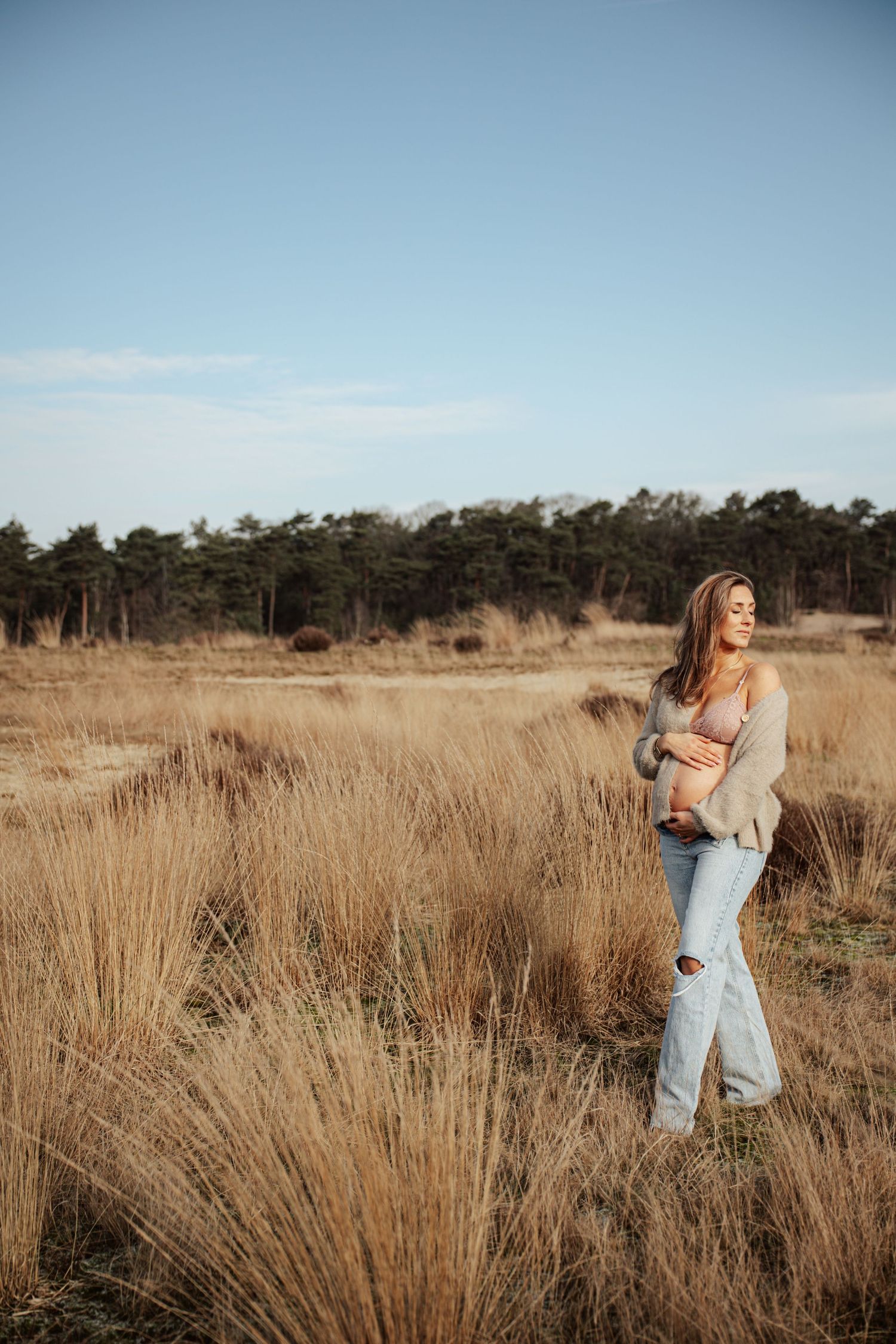 Casual maternity photo in golden grass field with pine trees in background.