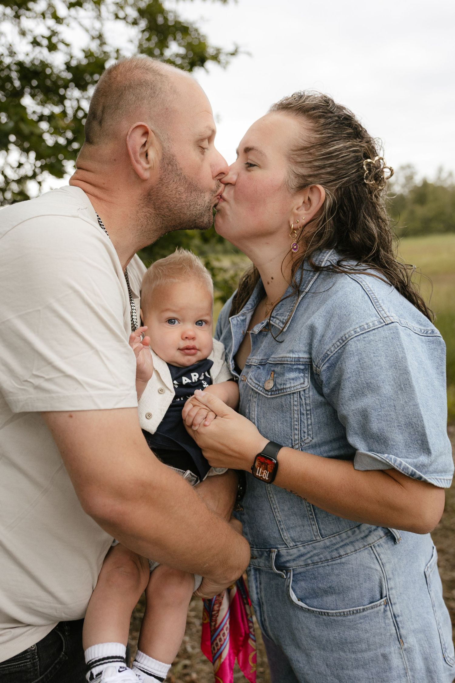 A couple kisses while holding their baby outdoors in a lush green setting, capturing a tender family moment.