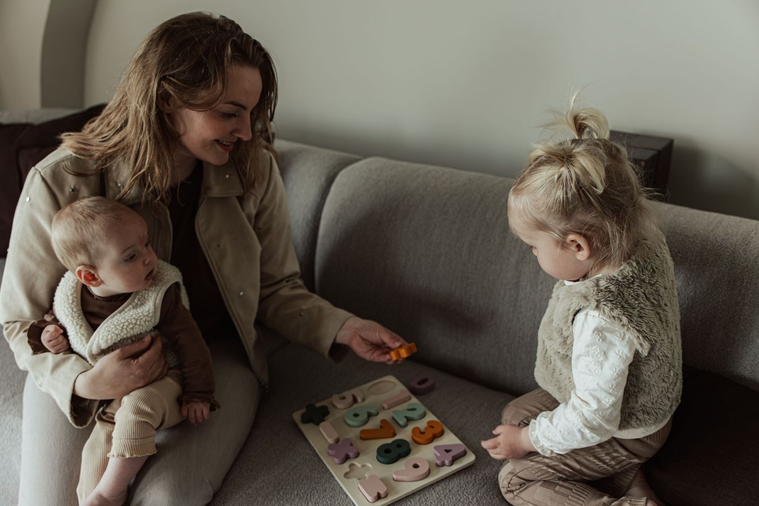 Mother sitting on sofa holding baby while toddler plays with colorful puzzle board.