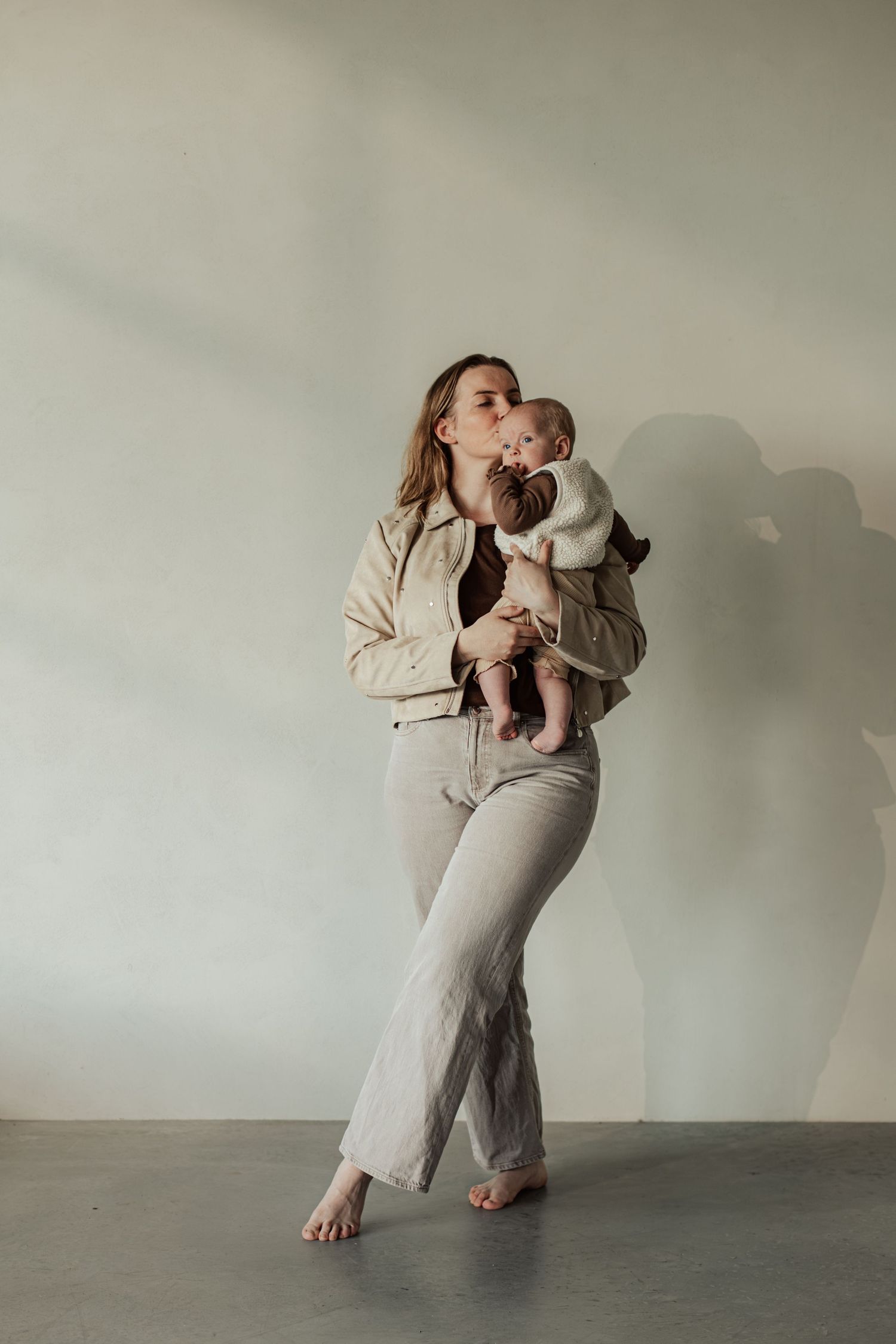 A barefoot mother in beige jeans and jacket kisses her newborn baby while standing against a light wall.
