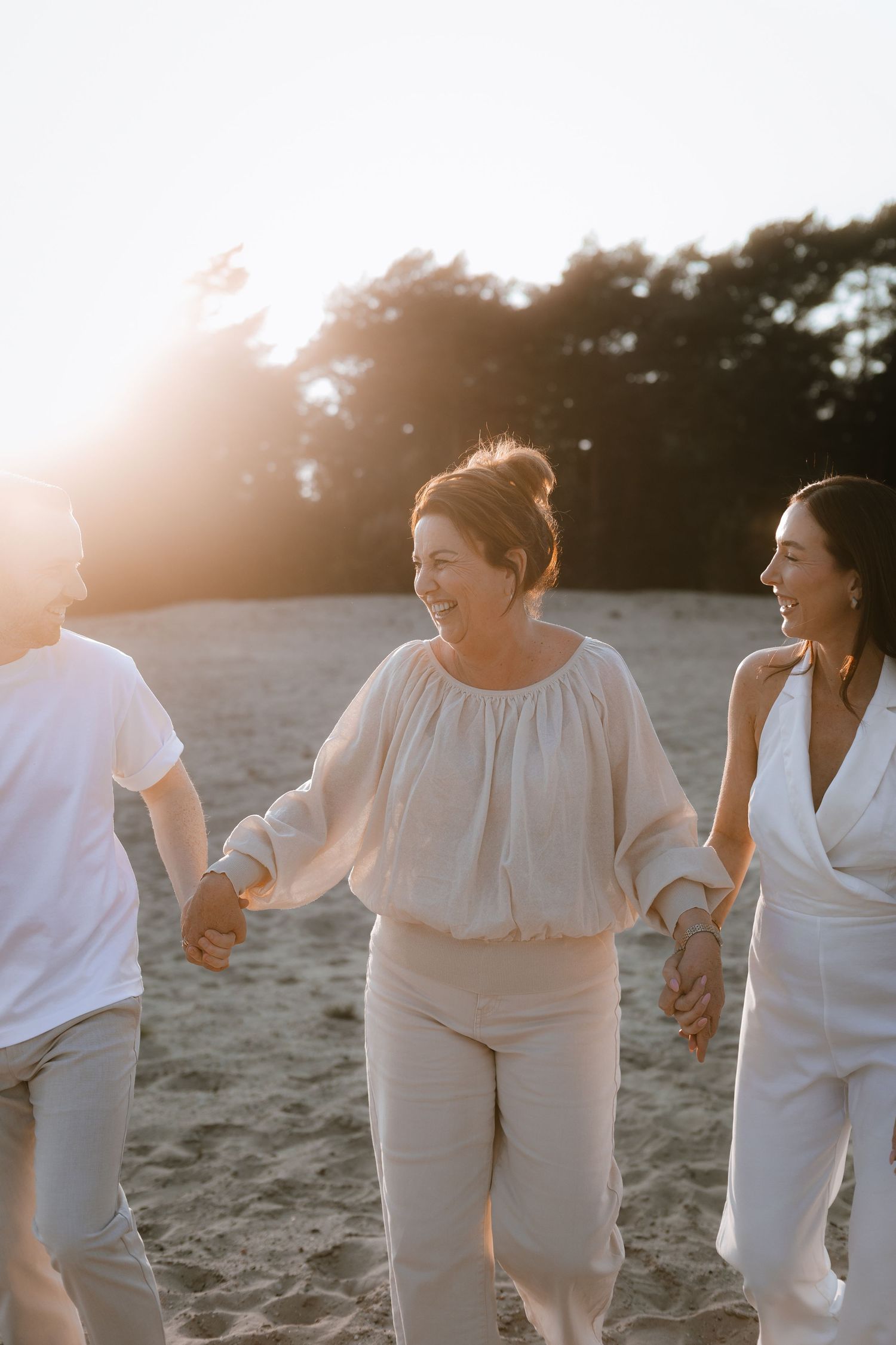 Three friends holding hands and laughing on a sandy beach at golden hour sunset.